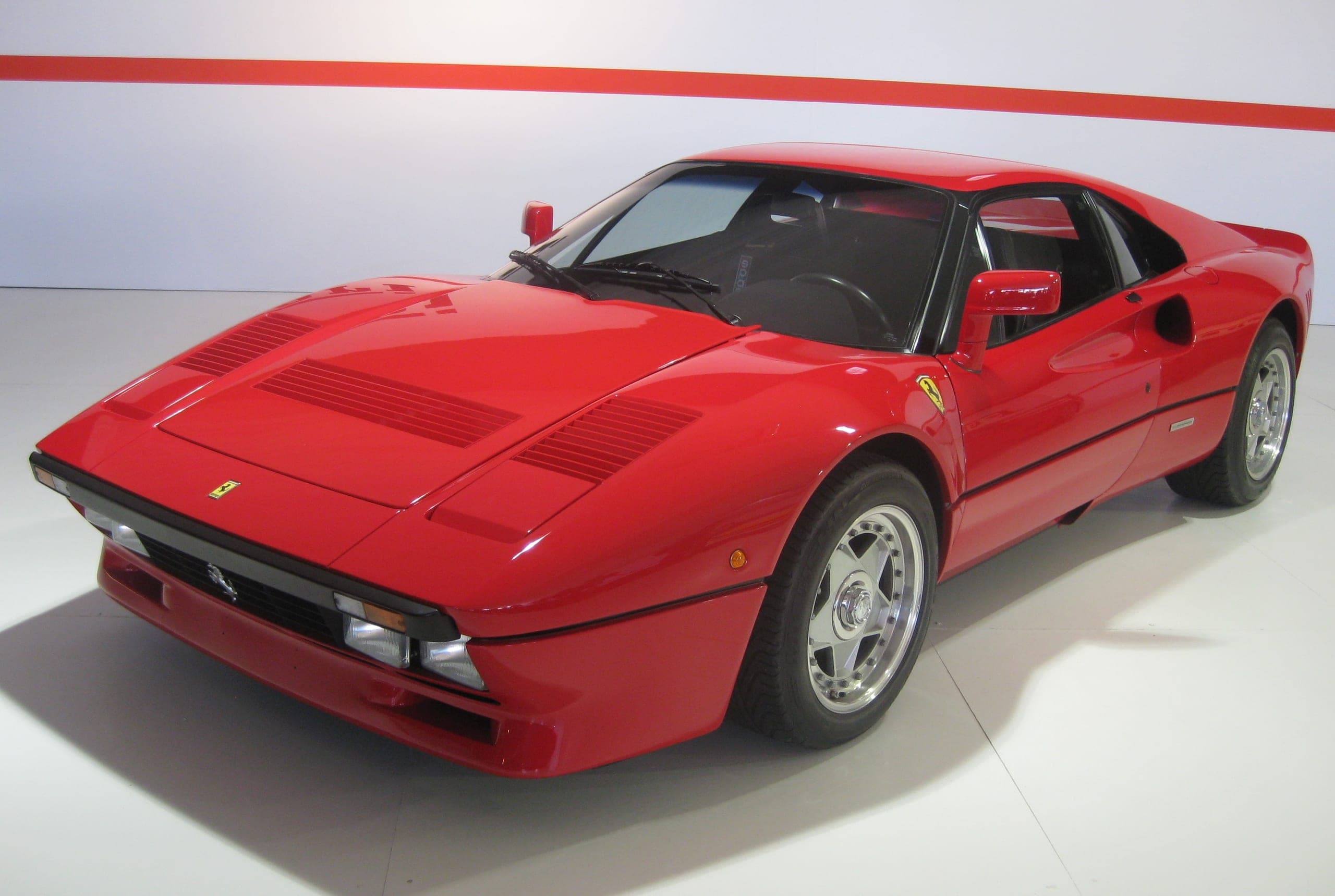 Ferrari 288 GTO roja sobre fondo blanco, vista de tres cuartos delantera con las tres branquias inclinadas tras el paso de rueda